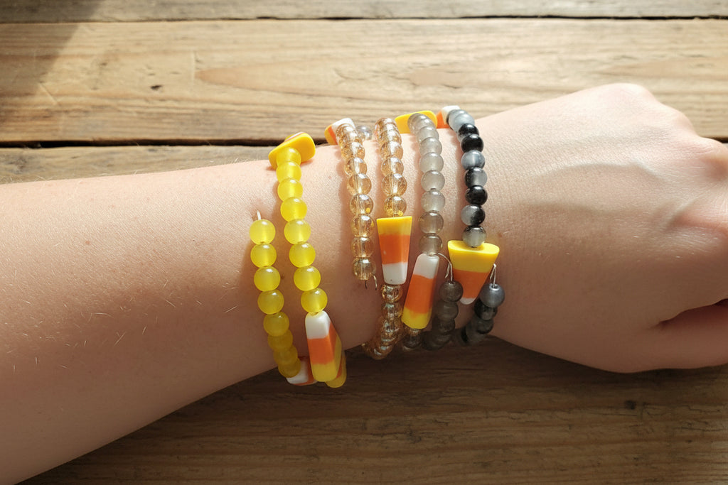 A close-up of a person's wrist wearing  beaded stretch bracelets, including yellow beads, clear beads, gray beads, and candy corn accents.