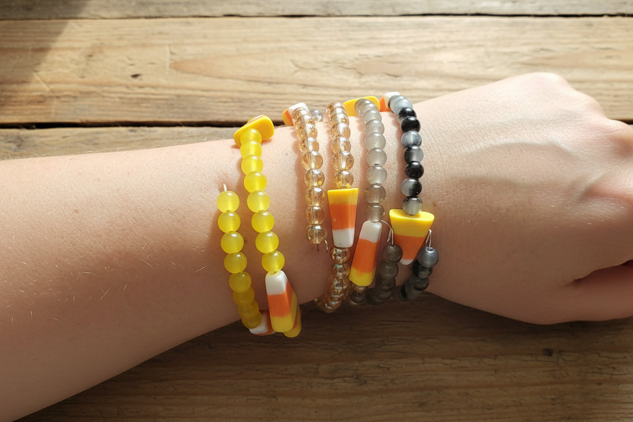 A close-up of a person's wrist wearing  beaded stretch bracelets, including yellow beads, clear beads, gray beads, and candy corn accents.