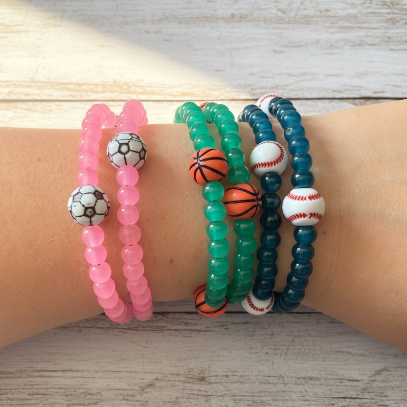 A stack of four beaded stretch bracelets on a person's wrist, with soccer, basketball, and baseball motifs.