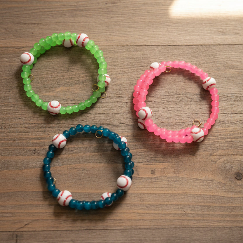Three beaded stretch bracelets with baseball charms, shown in green, pink, and blue on a dark surface.
