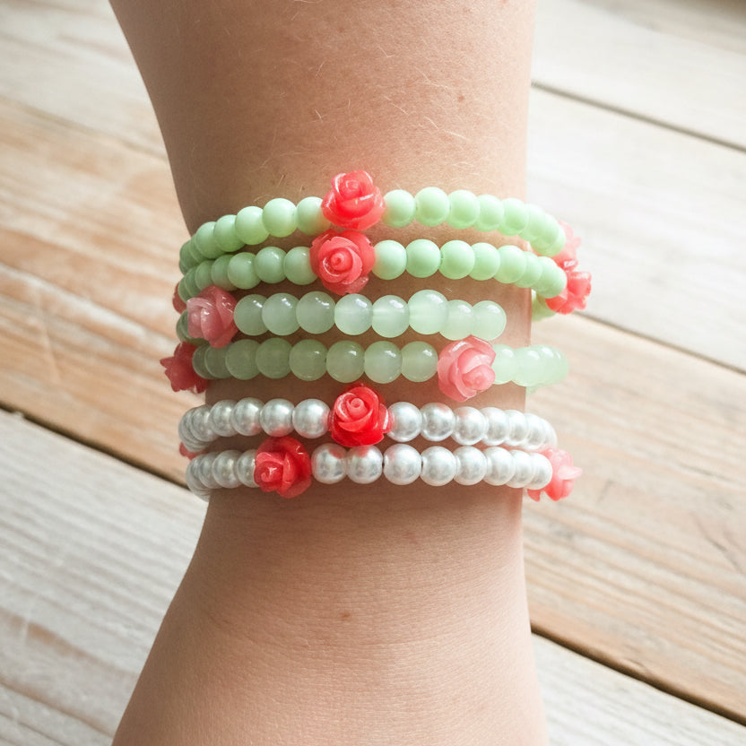 A stack of  beaded stretch bracelets worn on a wrist. The bracelets include light green beads, white faux pearl beads, and coral pink rose-shaped beads.