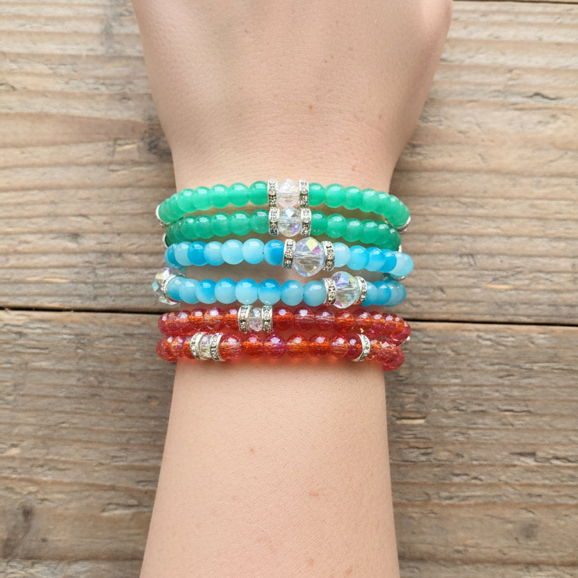 beaded stretch bracelets in green, light blue, and red, each with a clear rhinestone spacer, shown stacked on a person's wrist 