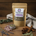 A stand-up kraft paper pouch labeled 'Astral Travel Incense' with a clear window showing dried herbs inside, A small open silver tin filled with the incense is placed in front, along with scattered pink crystal stones.