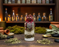 A small clear glass bottle filled with layered ingredients, topped with dark red wax and a red cord, with a silver heart-shaped ornament hanging from the neck. 