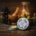 A tin of herbal balm with its lid off, displayed on a rustic wooden table surrounded by lavender, glass bottles, a wooden mortar and pestle, and warm fairy lights in the background. The label reads 'The Herbal Witch Co.