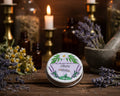 A silver tin of herbal wax balm labeled 'Sleep' with a lavender and green botanical illustration, shown next to its open lid containing pale yellow wax and a few green stones, with a glass bottle and fairy lights in the background.