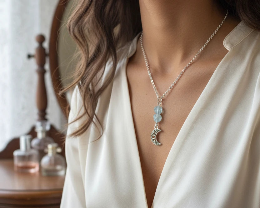 A silver chain necklace , featuring a dangling pendant with two light blue beads and a silver filigree crescent moon charm.