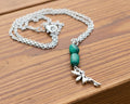 A silver chain necklace with two green beads and a silver fairy charm, displayed on a weathered wooden surface