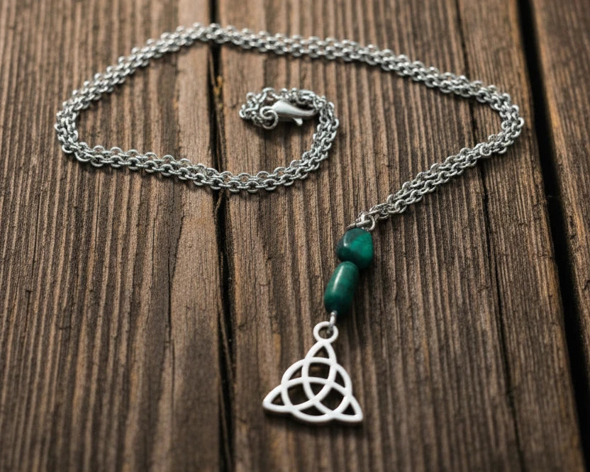 A silver Celtic knot triquetra pendant necklace with two green beads, displayed on a weathered wooden surface 