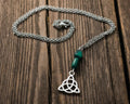 A silver Celtic knot triquetra pendant necklace with two green beads, displayed on a weathered wooden surface 