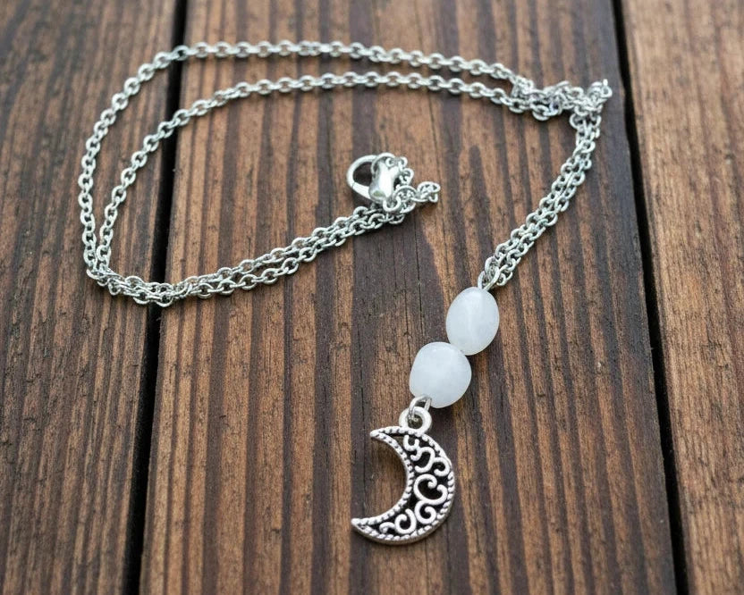 A silver chain necklace with a crescent moon pendant and two pale round beads, displayed on a weathered wooden surface 