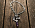 A silver tree of life pendant necklace with a dangling purple amethyst bead, displayed on a weathered wooden surface 