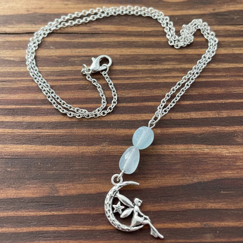 A silver-toned chain necklace with a fairy perched on a crescent moon and star charm, accented by two light blue oval beads, displayed on a weathered wooden surface 