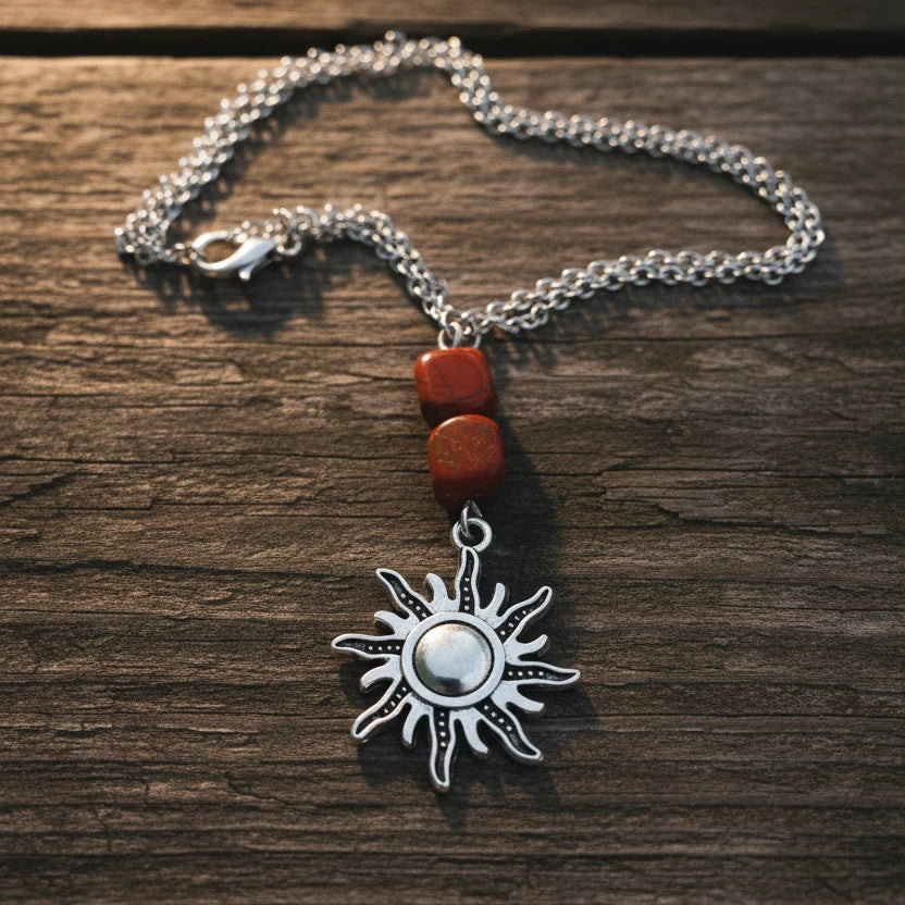 A silver-toned necklace featuring a sun pendant and two red beads, displayed on a weathered wooden surface 